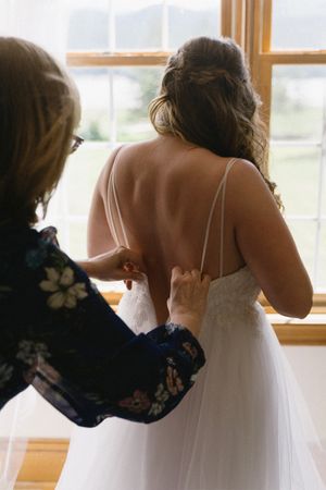 Behind the scenes photo of bride getting dressed by window on wedding day.