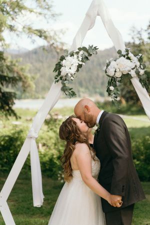 Bride and groom kiss under white floral arch with mountain backdrop.