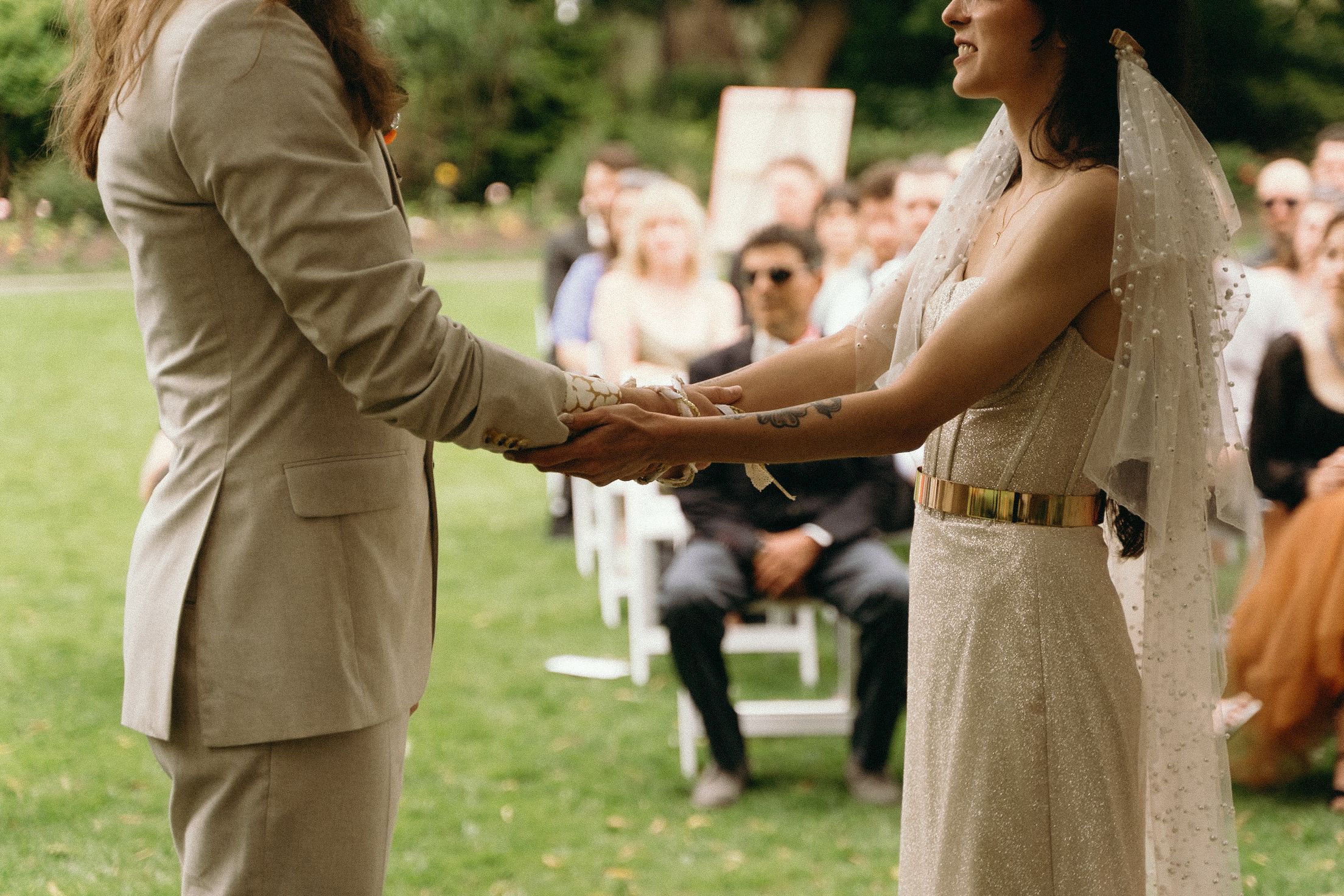 A couple holds hands during an outdoor wedding ceremony with guests seated on the lawn.