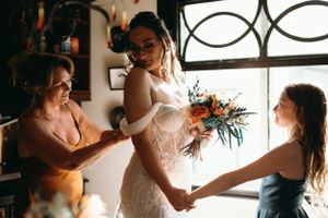 A bride gets help with final wedding dress preparations while holding a colorful bouquet.