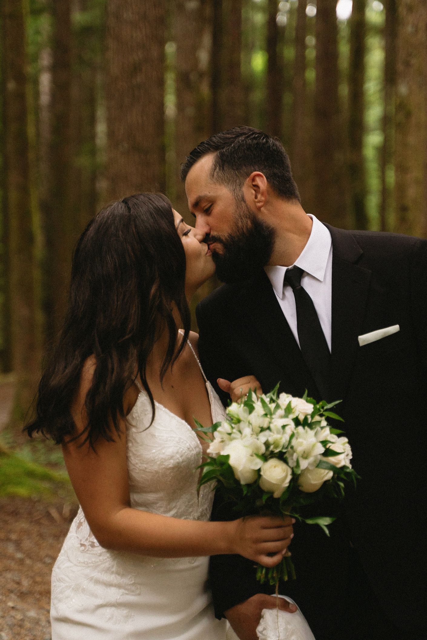 A romantic wedding couple shares a kiss in a forest setting while holding a white floral bouquet.