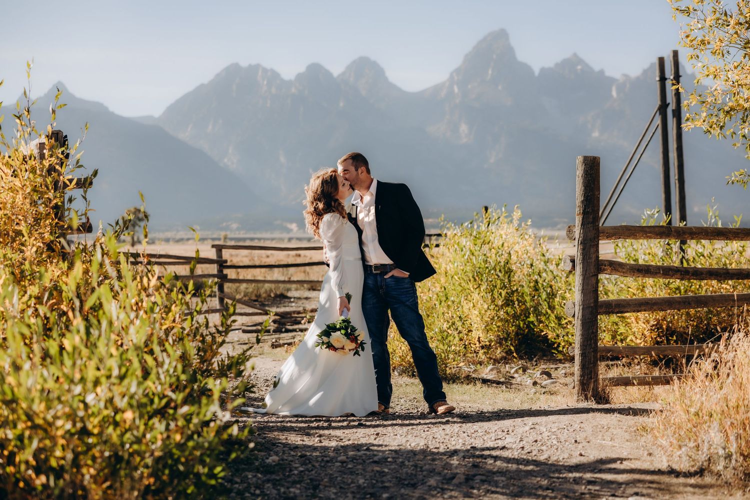 A romantic wedding couple poses near rustic wooden structures in a desert landscape with mountains in the background.