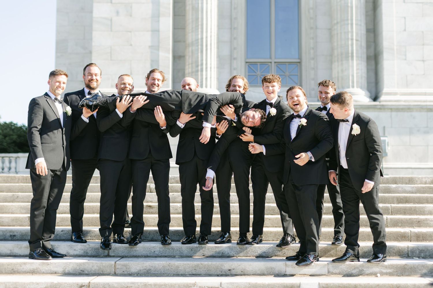 Groomsmen show off their colorful socks while sitting on stone steps outside an elegant building.