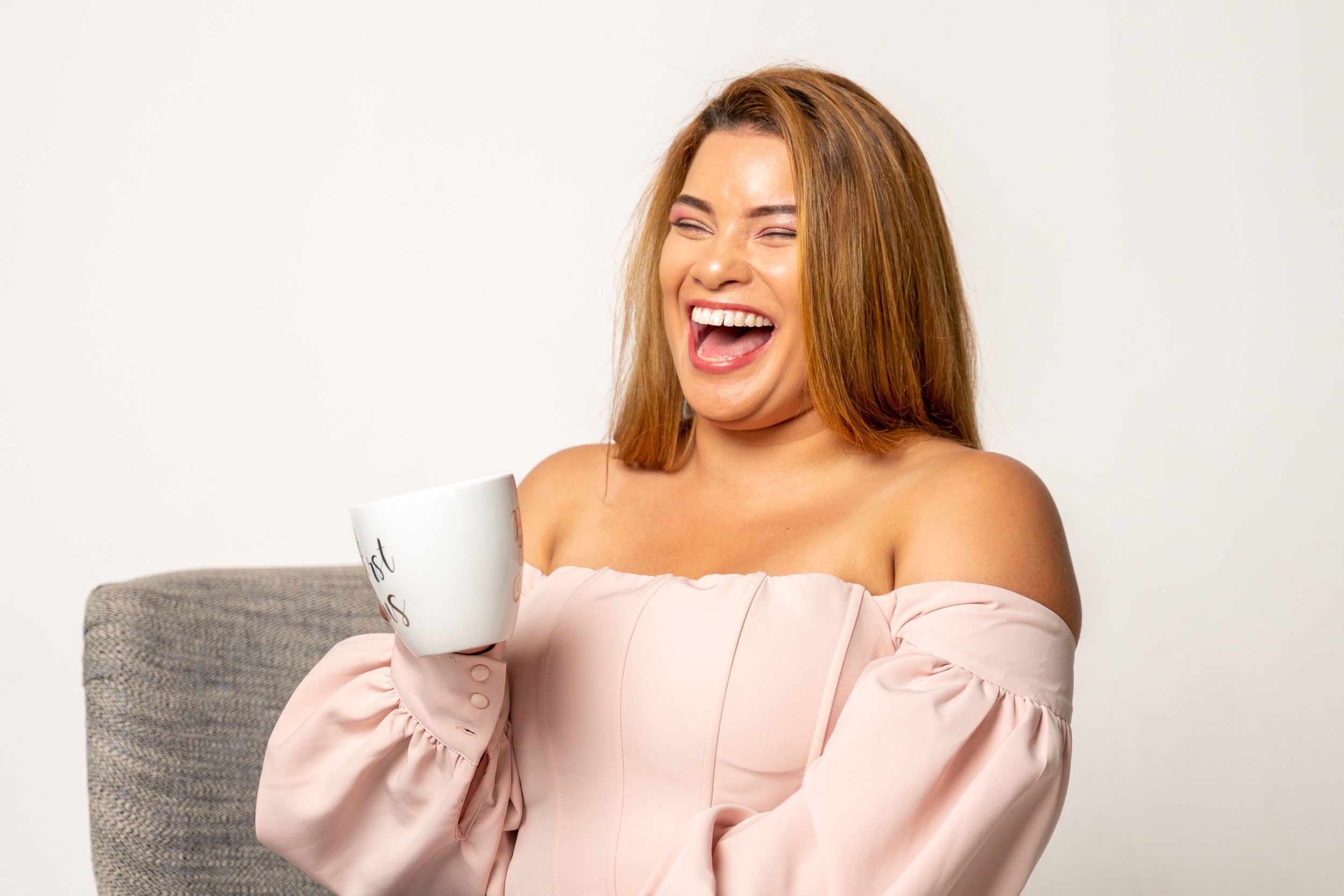 Woman wearing pink off-shoulder top laughs joyfully while sitting on grey couch.