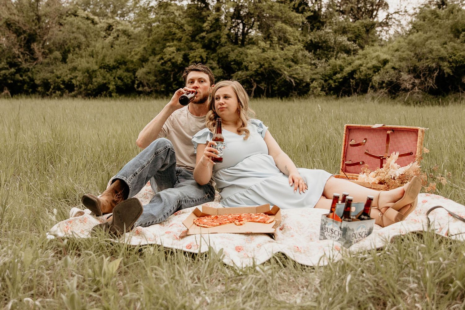 A couple enjoys a romantic outdoor picnic on a white blanket in a grassy meadow during golden hour.