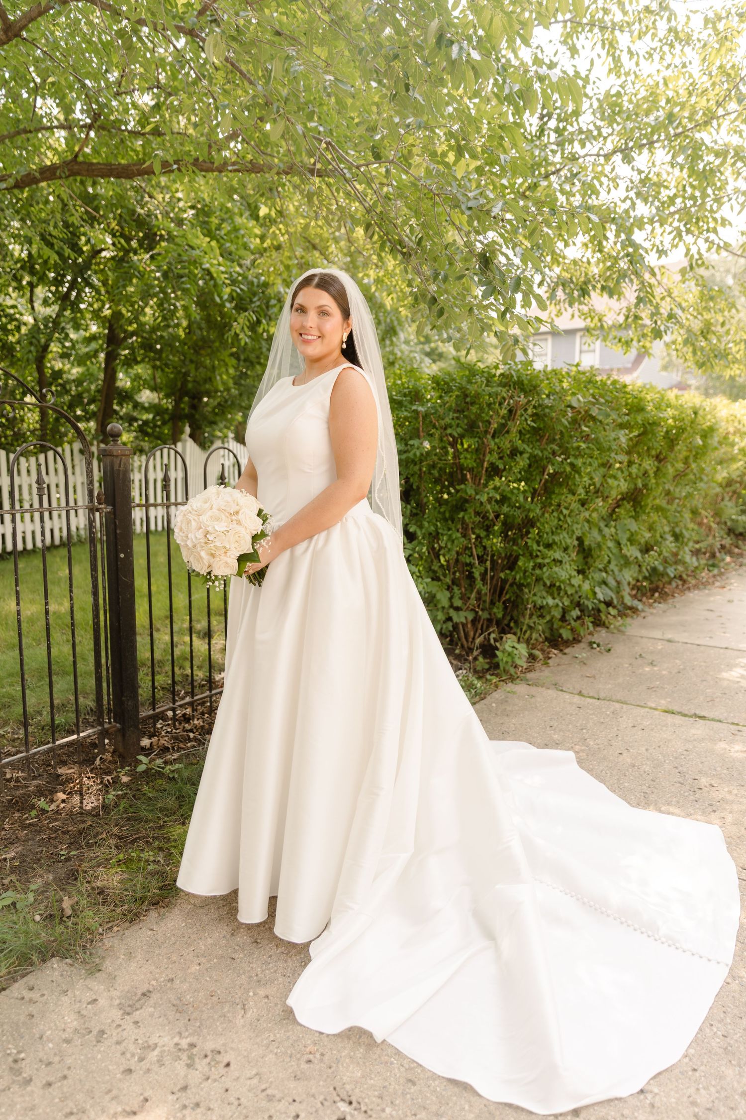 A bride in an elegant white satin gown with a long train poses in a garden setting holding a white floral bouquet.
