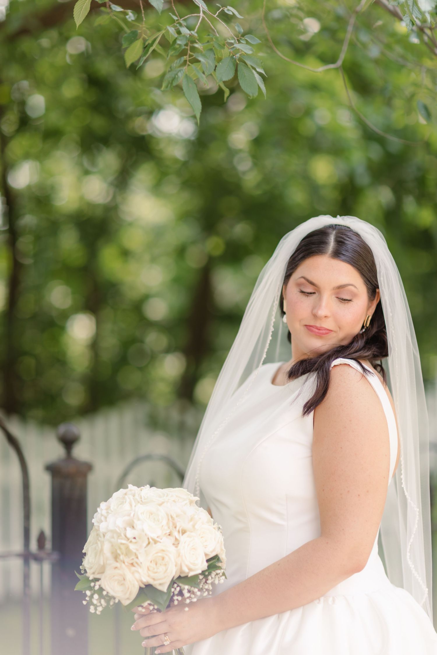 A bride in a white wedding dress and veil holds a bouquet of white flowers in an outdoor garden setting.