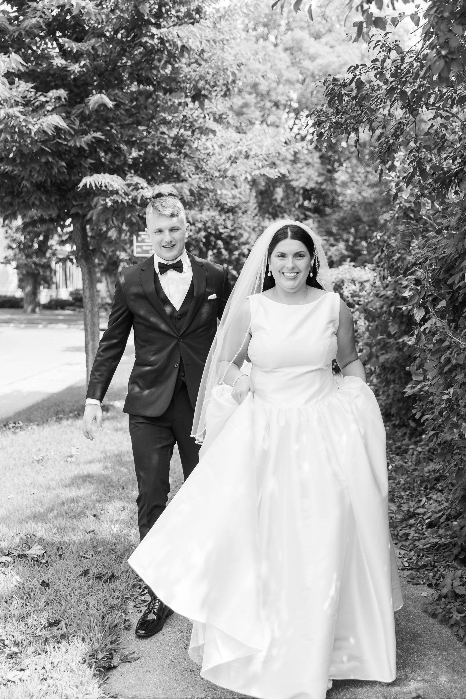 A black and white wedding photo showing a couple walking together outdoors along a tree-lined path.