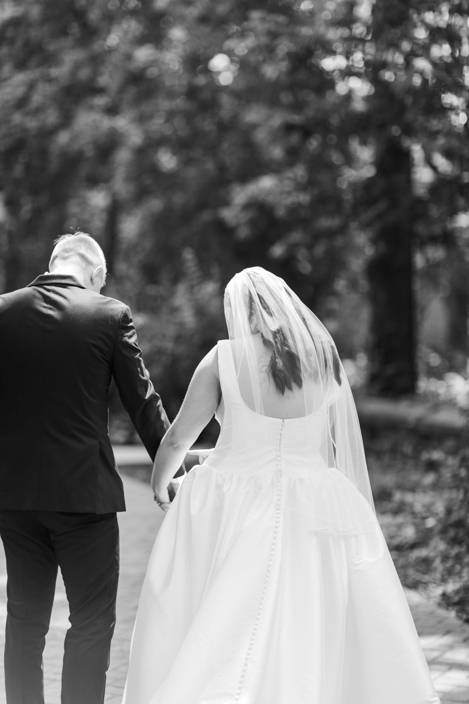 Black and white photo of a wedding party walking through a garden with the bride's flowing white dress and veil trailing behind.