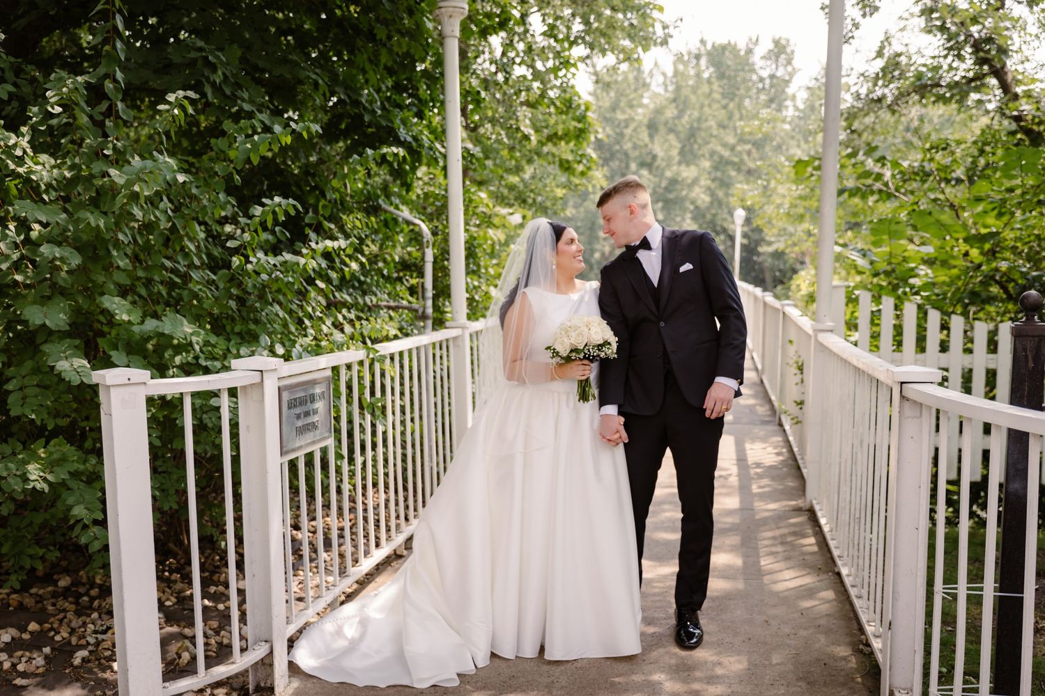 A couple in wedding attire walks together on a white railed bridge surrounded by lush green trees.