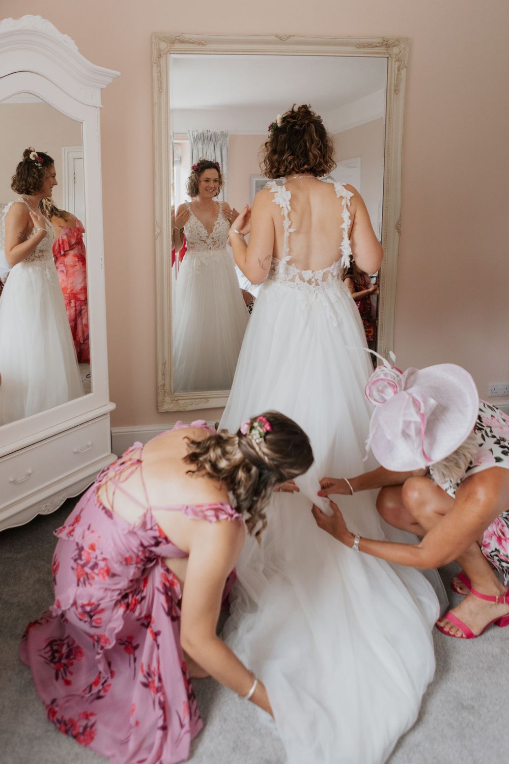Bridesmaids helping bride into her lace wedding gown in front of a large mirror in a pink bridal suite.