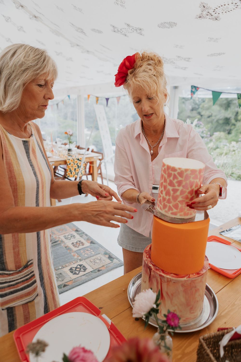 Two women assembling a vibrant orange and pink tiered wedding cake inside a decorated marquee tent.