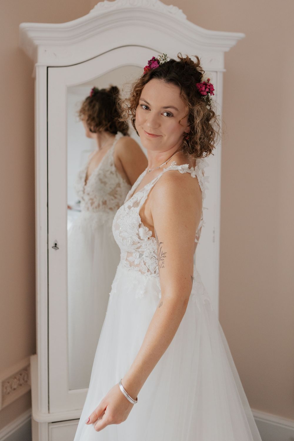Curly-haired bride in lace wedding dress with pink flowers in hair posing beside a white wardrobe mirror.