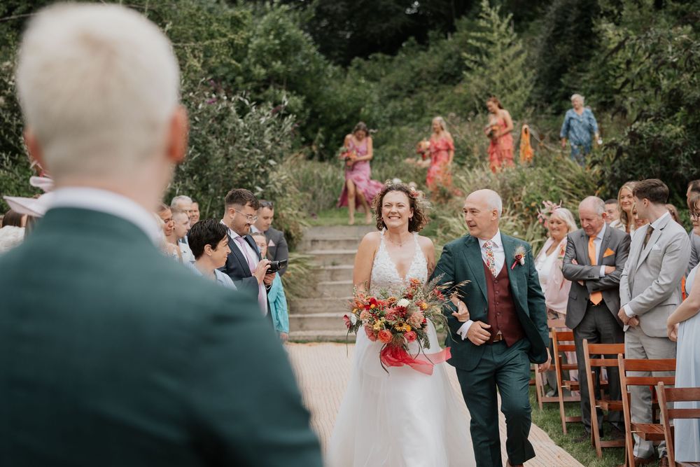 Bride walking down the outdoor aisle with her father past seated wedding guests in a garden ceremony.