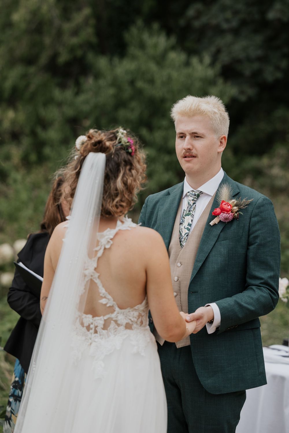 Groom in green suit holding hands with bride during outdoor wedding ceremony beneath a rustic arch.
