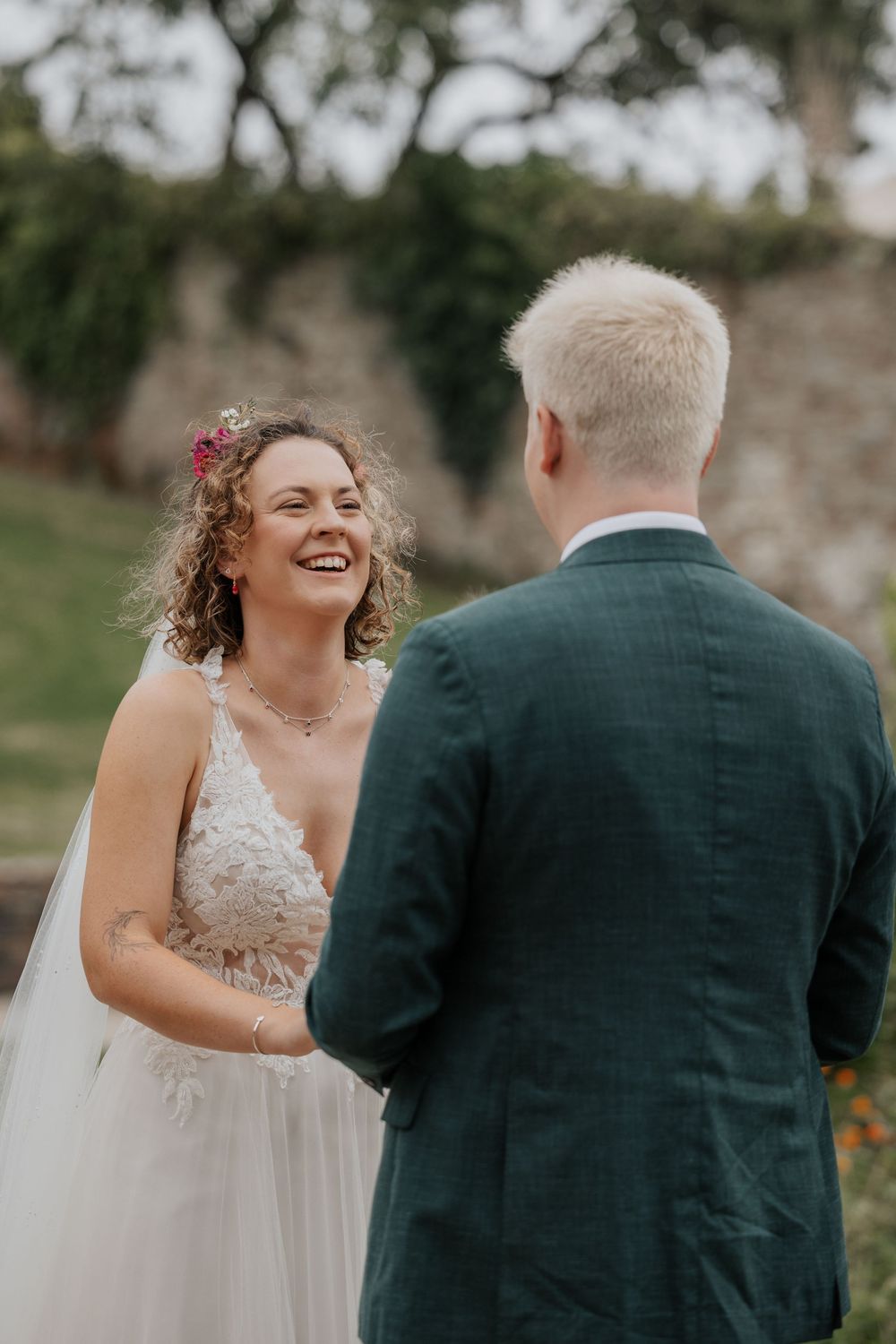 Bride laughing joyfully during outdoor wedding ceremony while holding hands with groom in dark green suit.