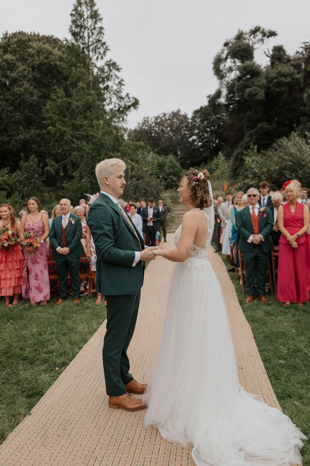 Bride and groom exchanging vows at outdoor wedding ceremony with guests standing in background.