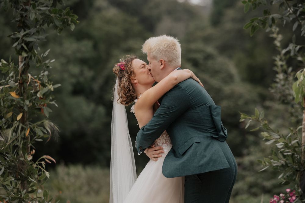 Bride and groom sharing first kiss surrounded by lush green foliage at outdoor wedding ceremony.