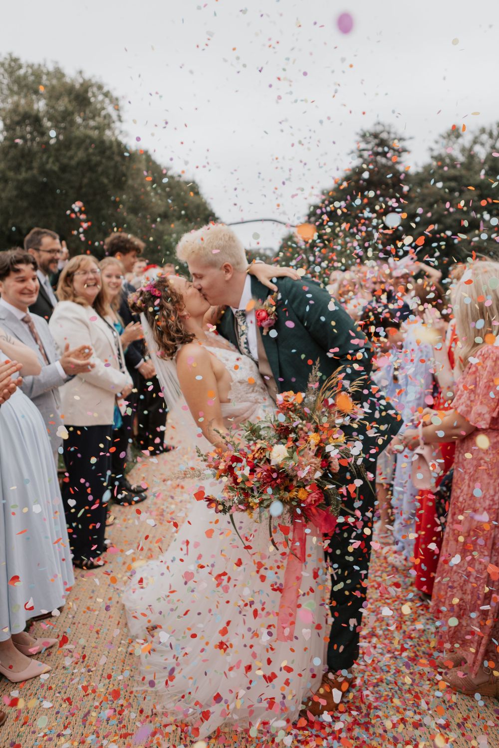 Bride and groom kissing surrounded by colorful confetti shower with wedding guests cheering.