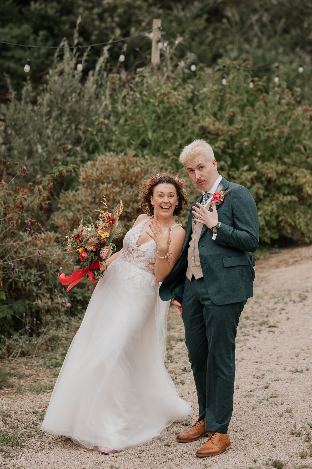 Laughing bride in lace gown and groom in green suit posing playfully in garden setting.
