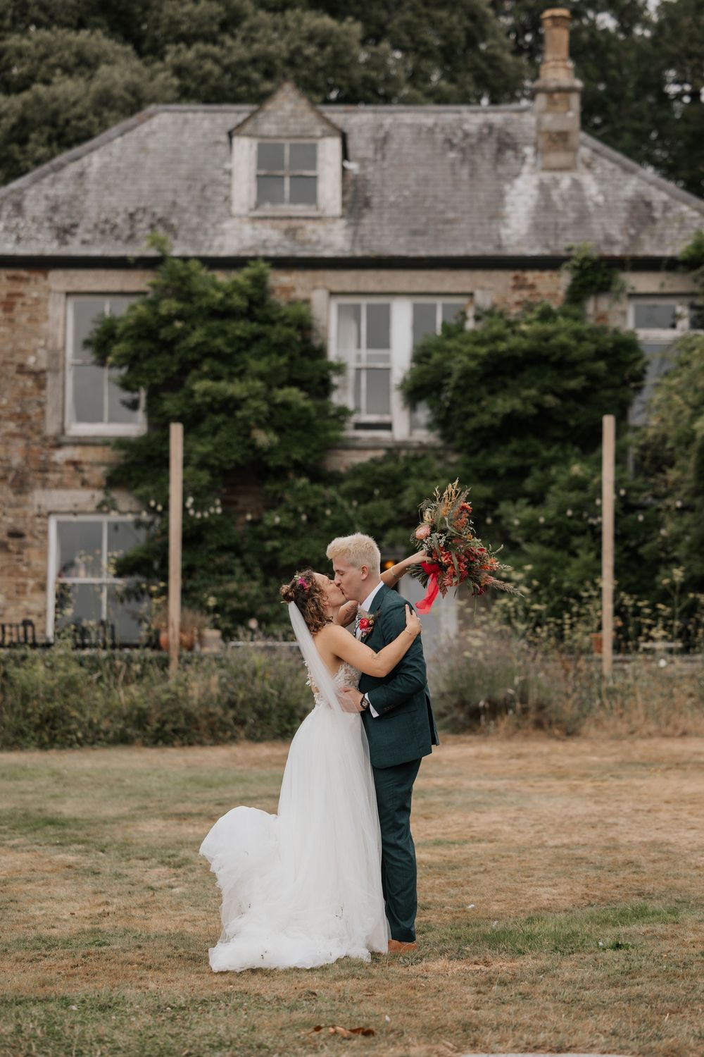 Bride and groom sharing romantic kiss on lawn in front of charming stone country house.