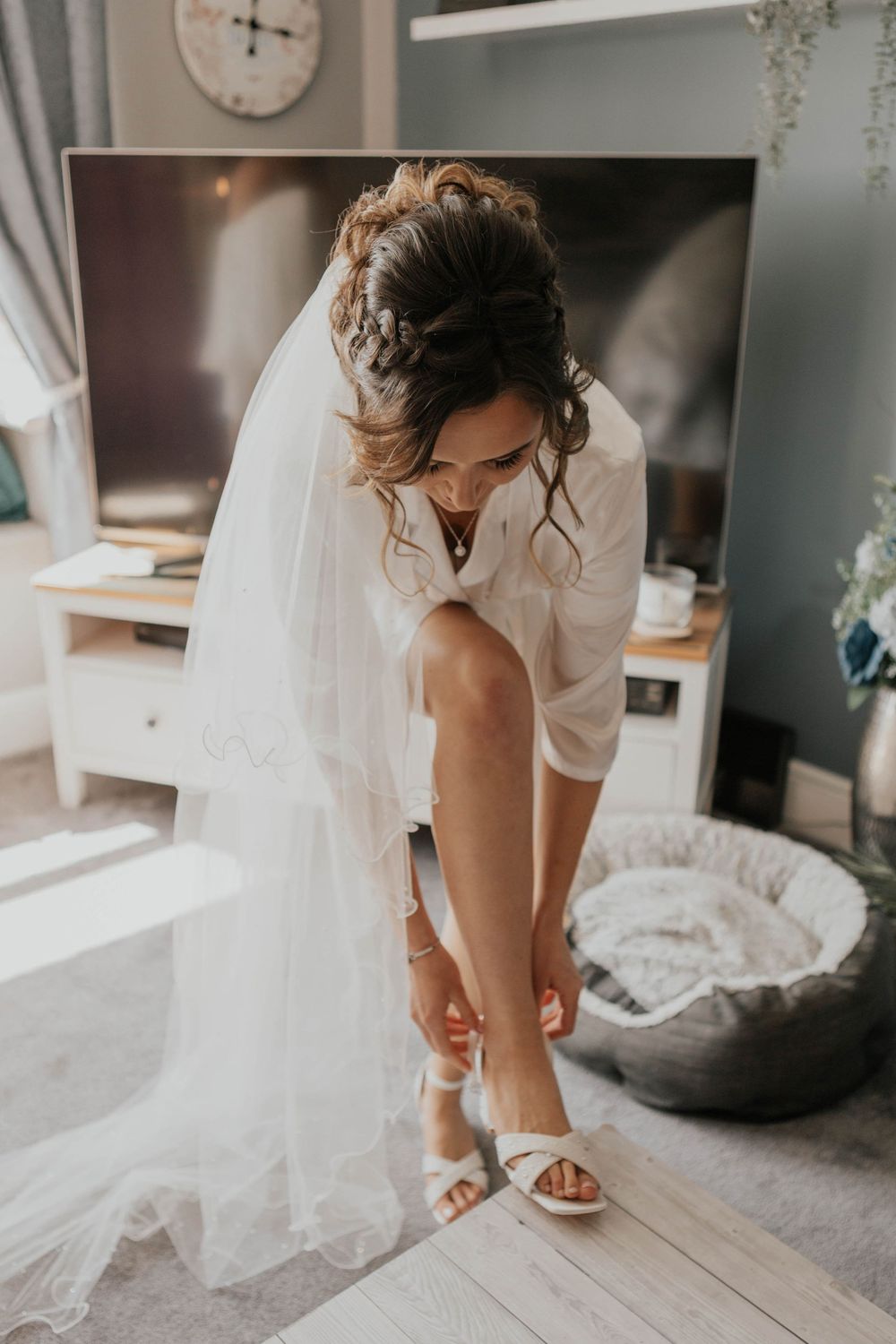 Bride in white shirt and veil fastens strappy white heels while getting ready for her wedding day.