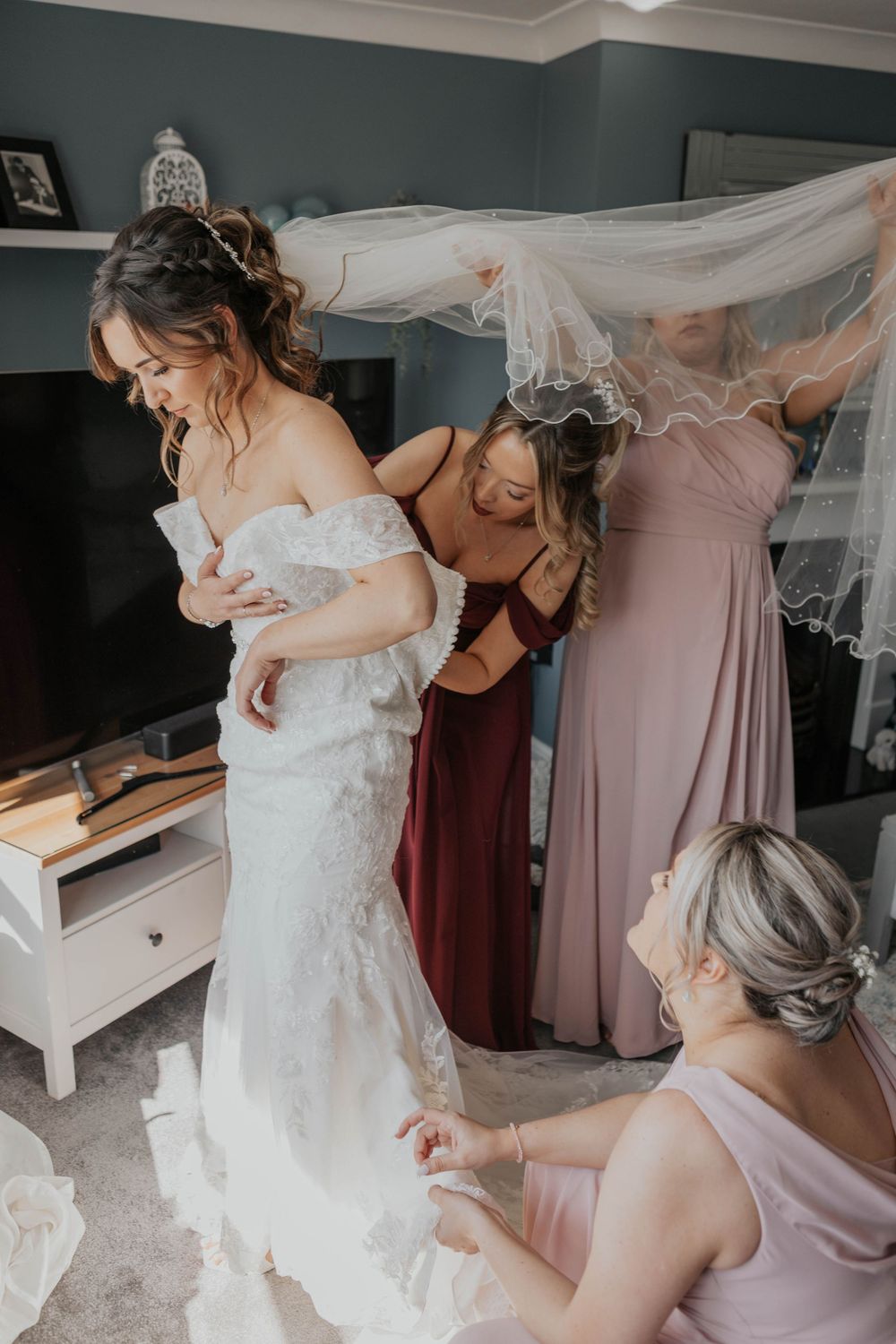 Bridesmaids in burgundy and pink gowns help bride into lace wedding dress and arrange flowing veil.