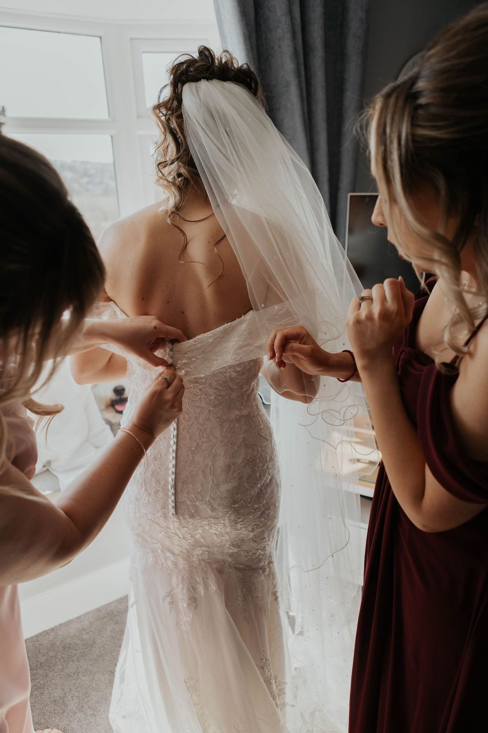 Two bridesmaids carefully fasten buttons on back of brides lace wedding gown by bright window.