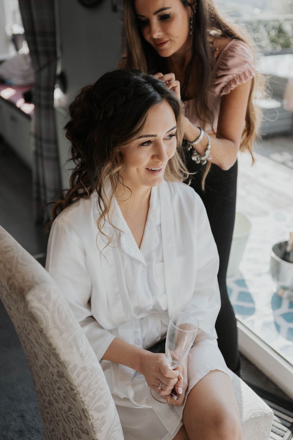 Smiling bride in white robe holds champagne glass while hairstylist works on her elegant updo.