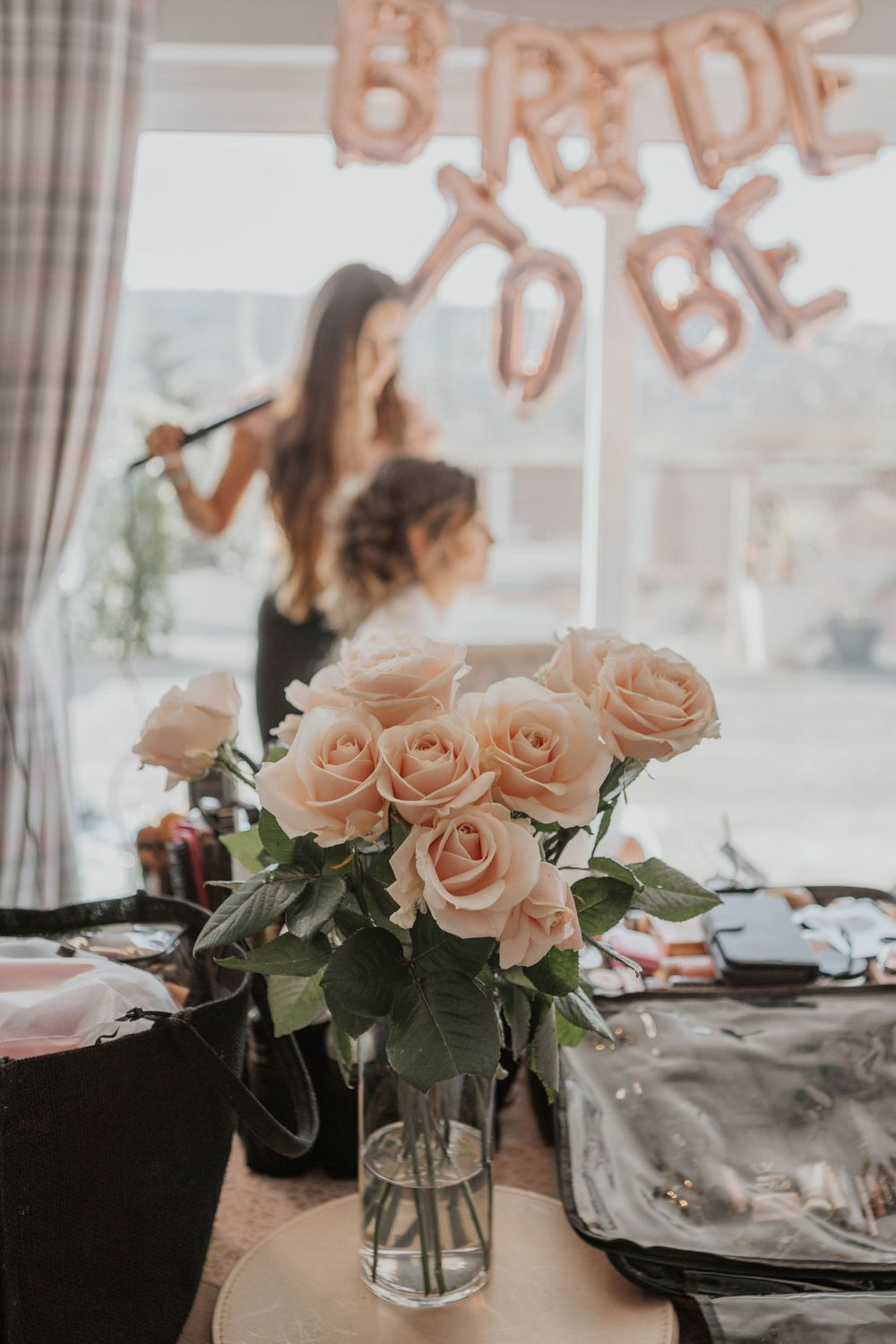 Bouquet of soft pink roses in glass vase with bride getting hair styled and BRIDE TO BE balloons behind.