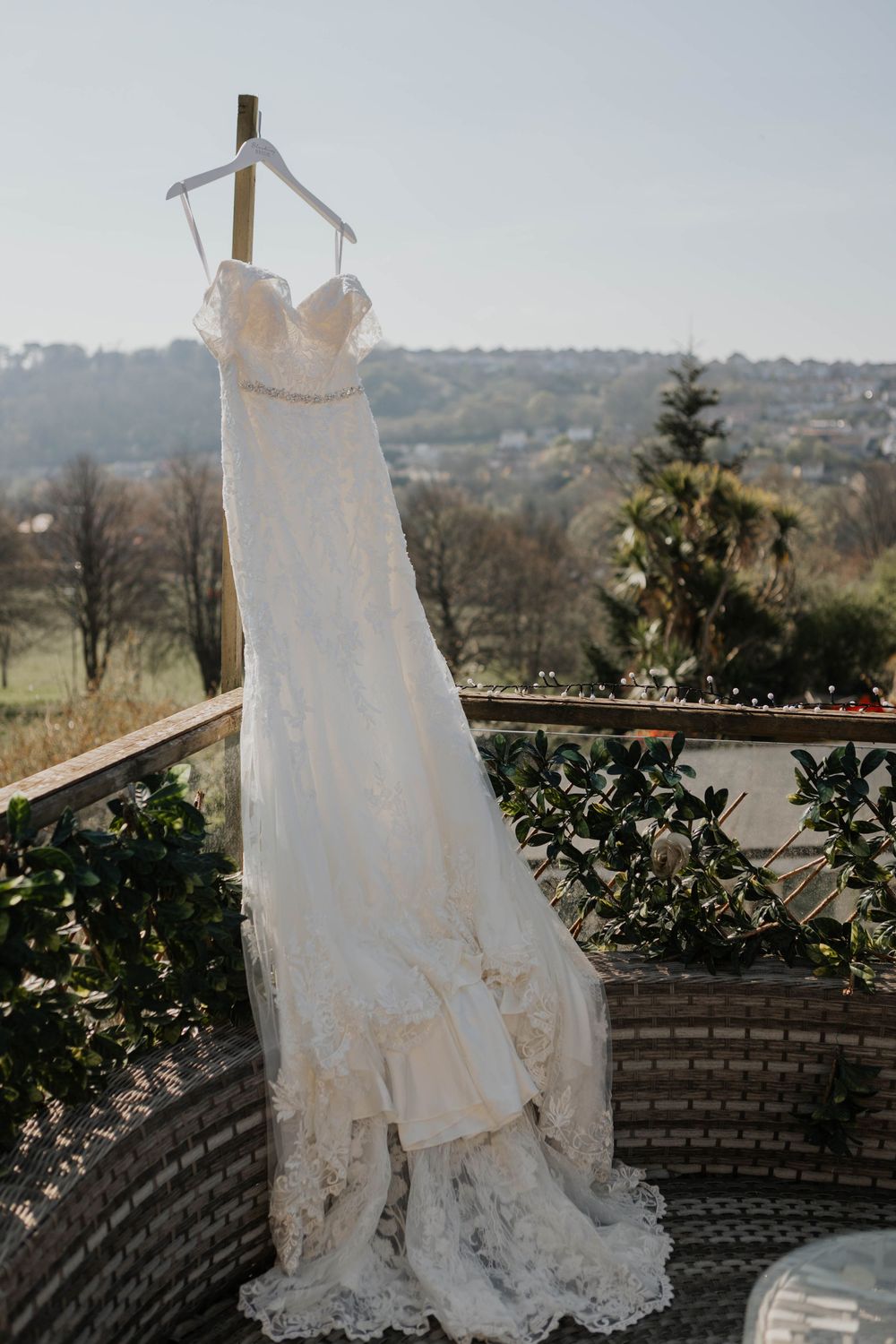 Elegant lace wedding dress hanging on balcony railing with scenic countryside landscape in background.