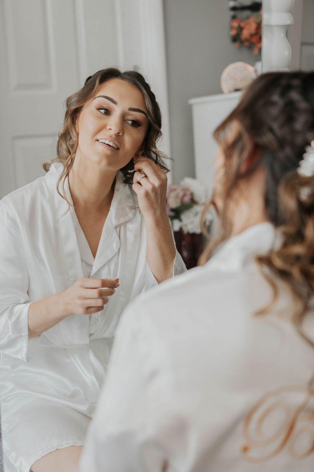 Beautiful bride in white robe smiles at mirror reflection while putting on earrings before wedding.