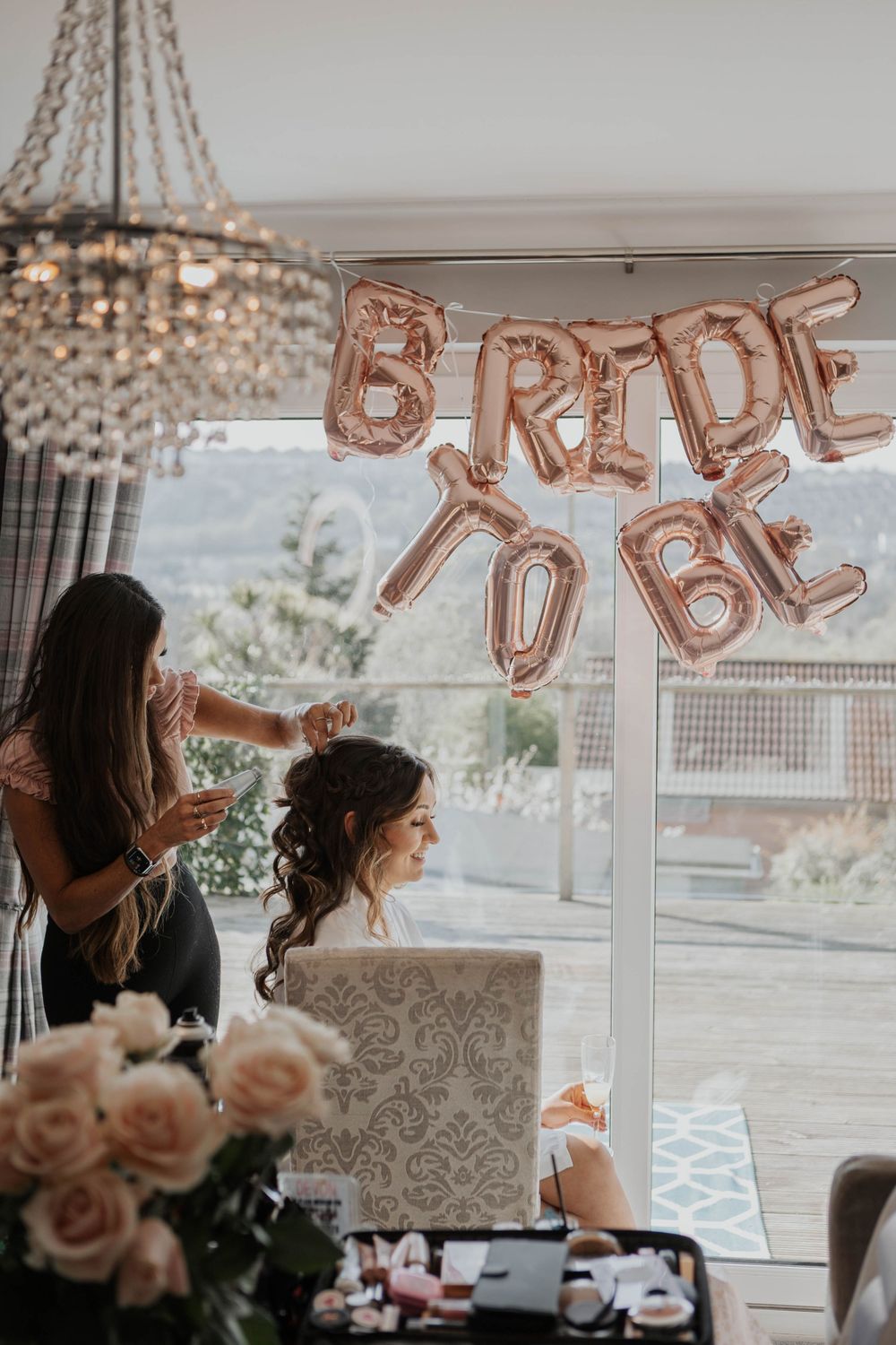 Bride-to-be gets hair styled under rose gold BRIDE TO BE balloons in a bright room with crystal chandelier.