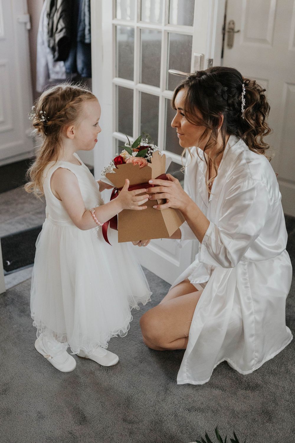 Bride in white satin robe kneels to receive gift box from adorable flower girl in white dress.