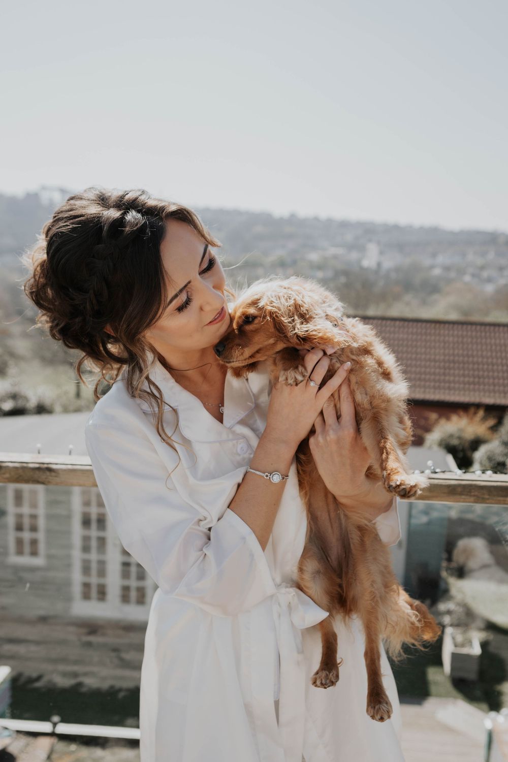 Bride in white robe tenderly kisses her small golden Cavalier King Charles Spaniel on sunny balcony.
