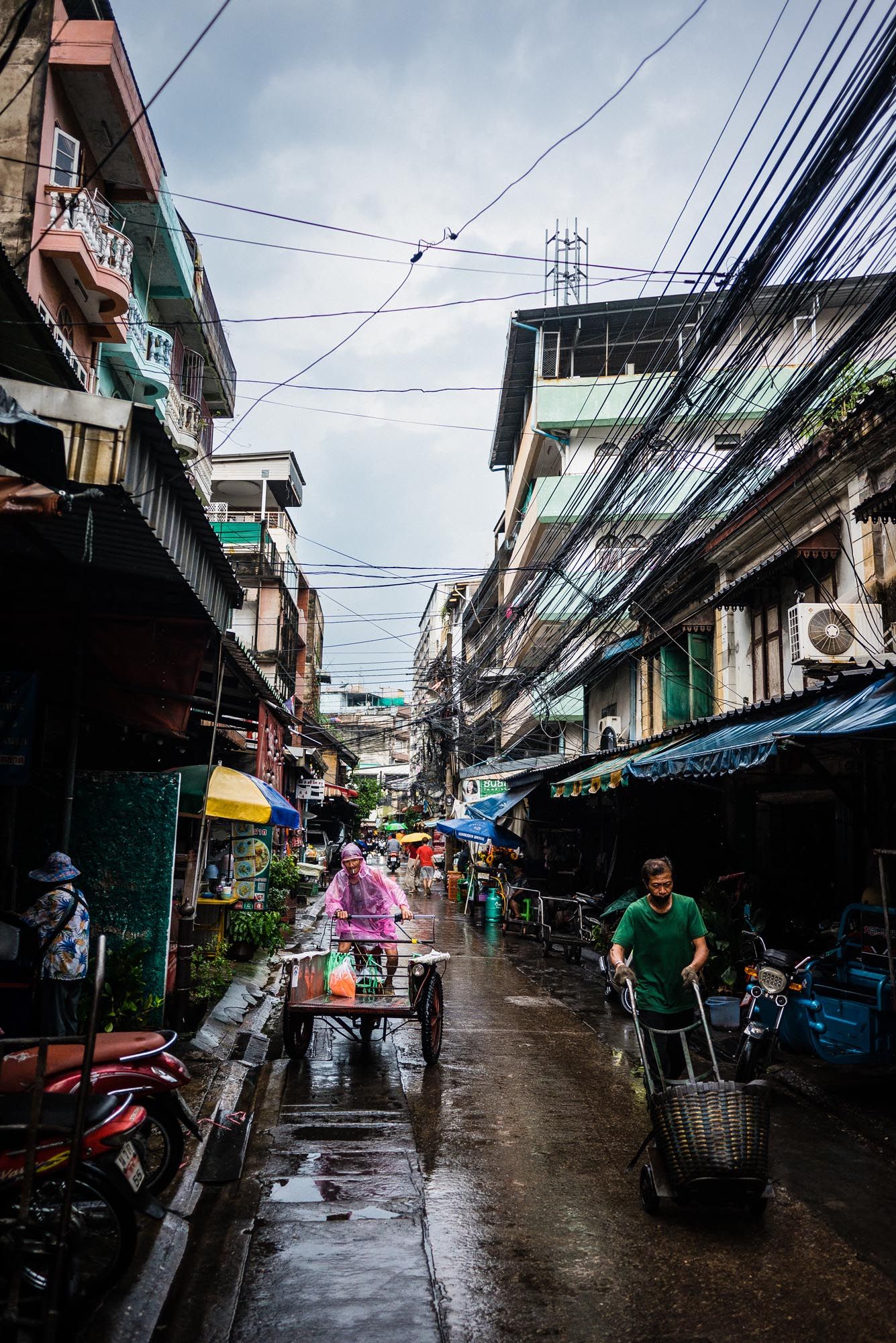 Narrow urban alleyway in Bangkok with power lines overhead and closed shops during a rainy day.