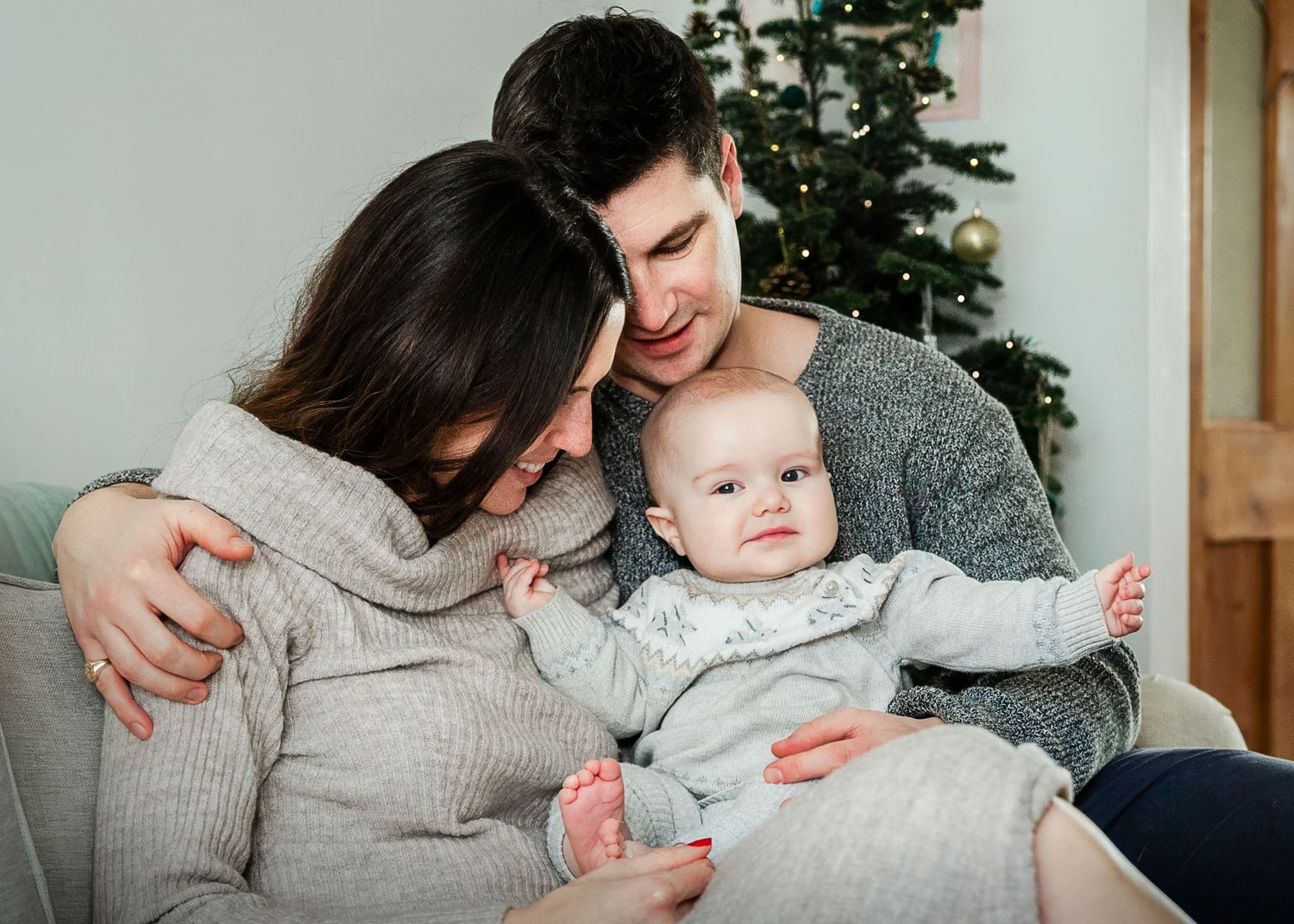 A baby sits with his mum and dad in front on the tree. They are hugging and look happy and relaxed during their Bristol Christmas photoshoot