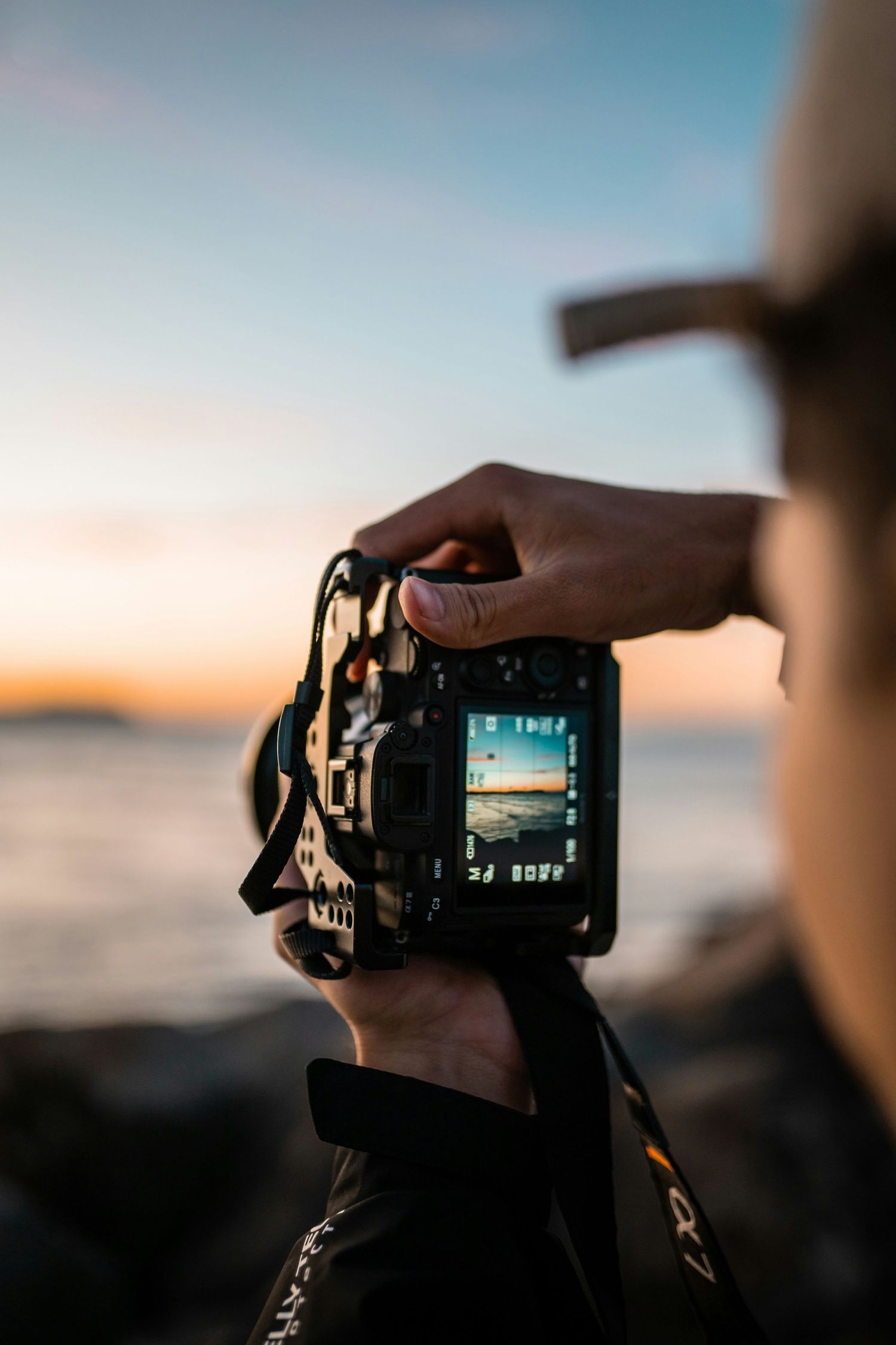 A hand holds a digital camera capturing a scenic sunset view through the LCD screen display.