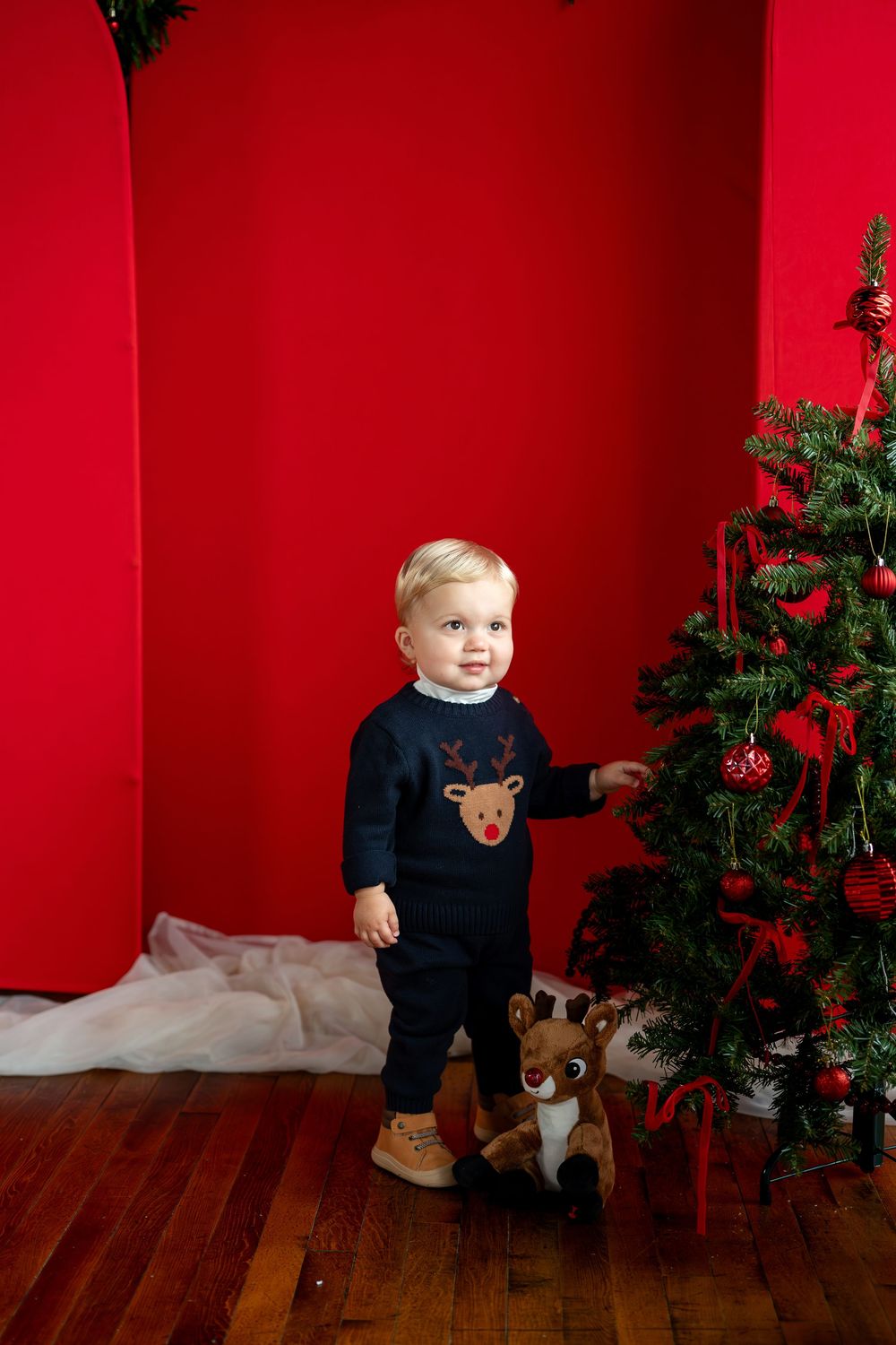 Toddler in navy reindeer outfit stands by decorated Christmas tree against red wall.