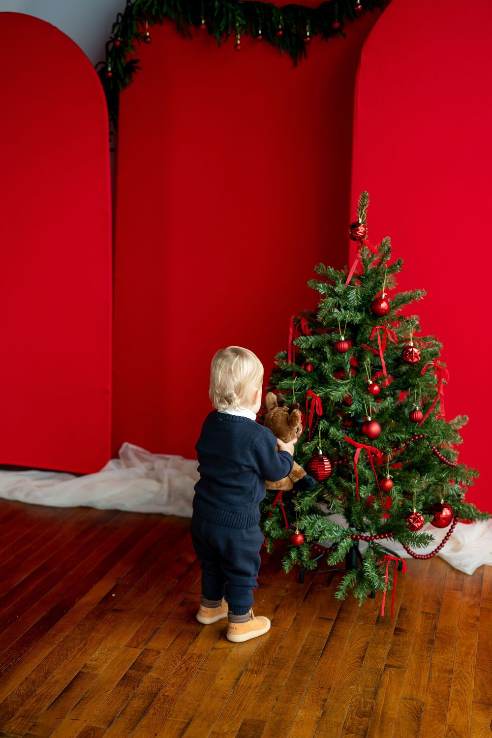 Baby in navy blue outfit standing with a teddy bear next to a Christmas tree with red decorations.