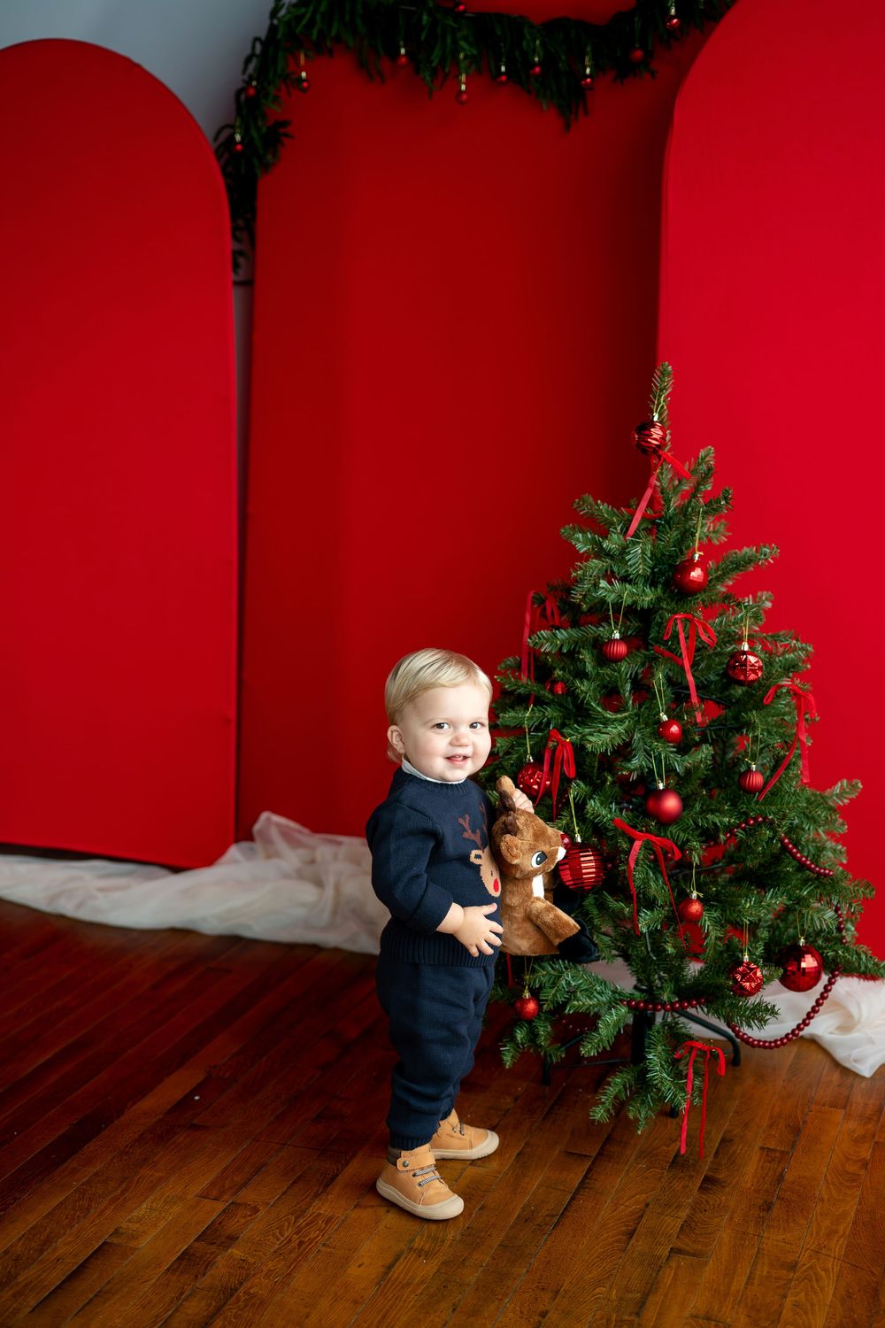 Happy toddler in navy reindeer sweater holding stuffed animal beside a Christmas tree with red decor.