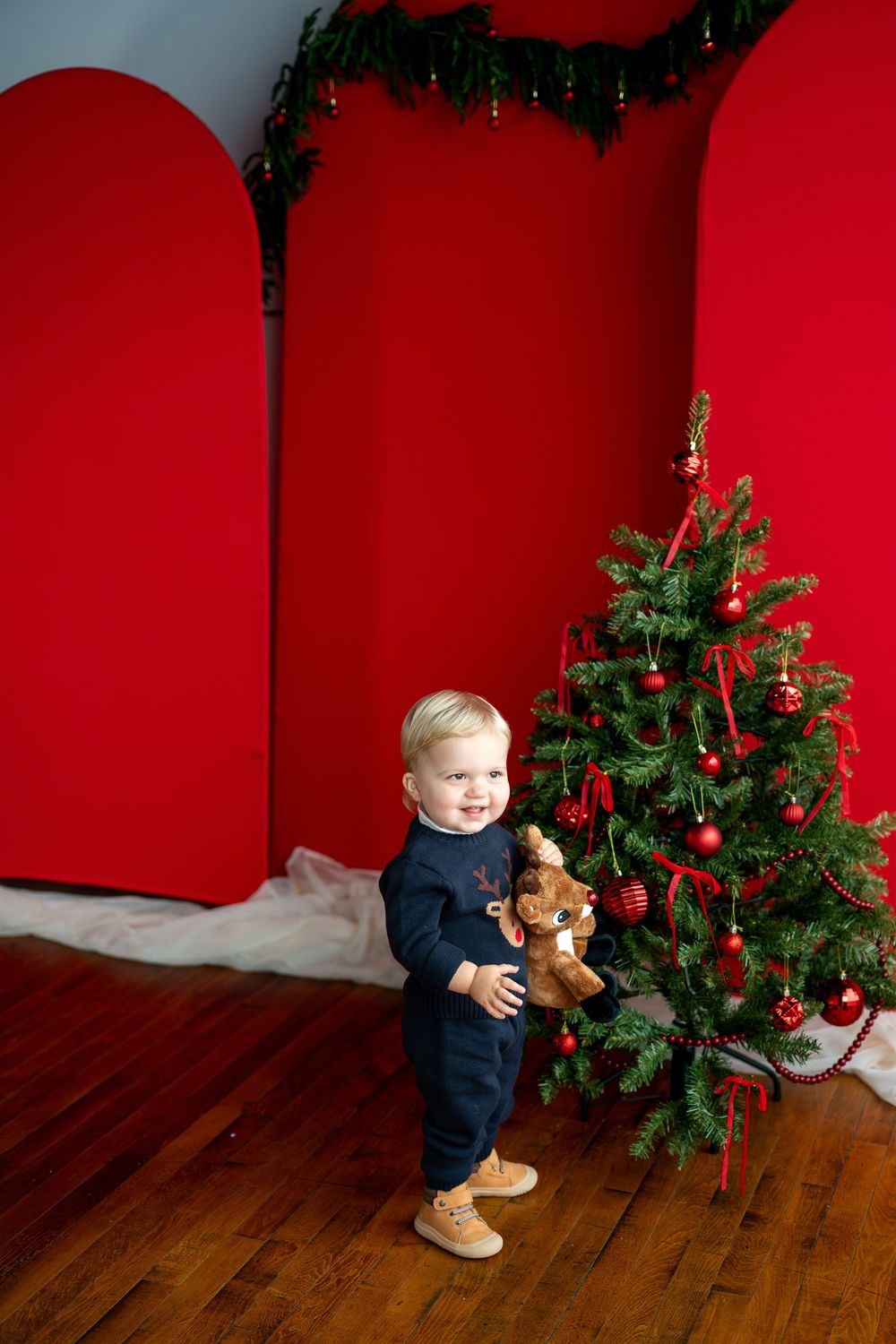Cheerful toddler in navy reindeer outfit smiling beside a Christmas tree with red ornaments and backdrop.