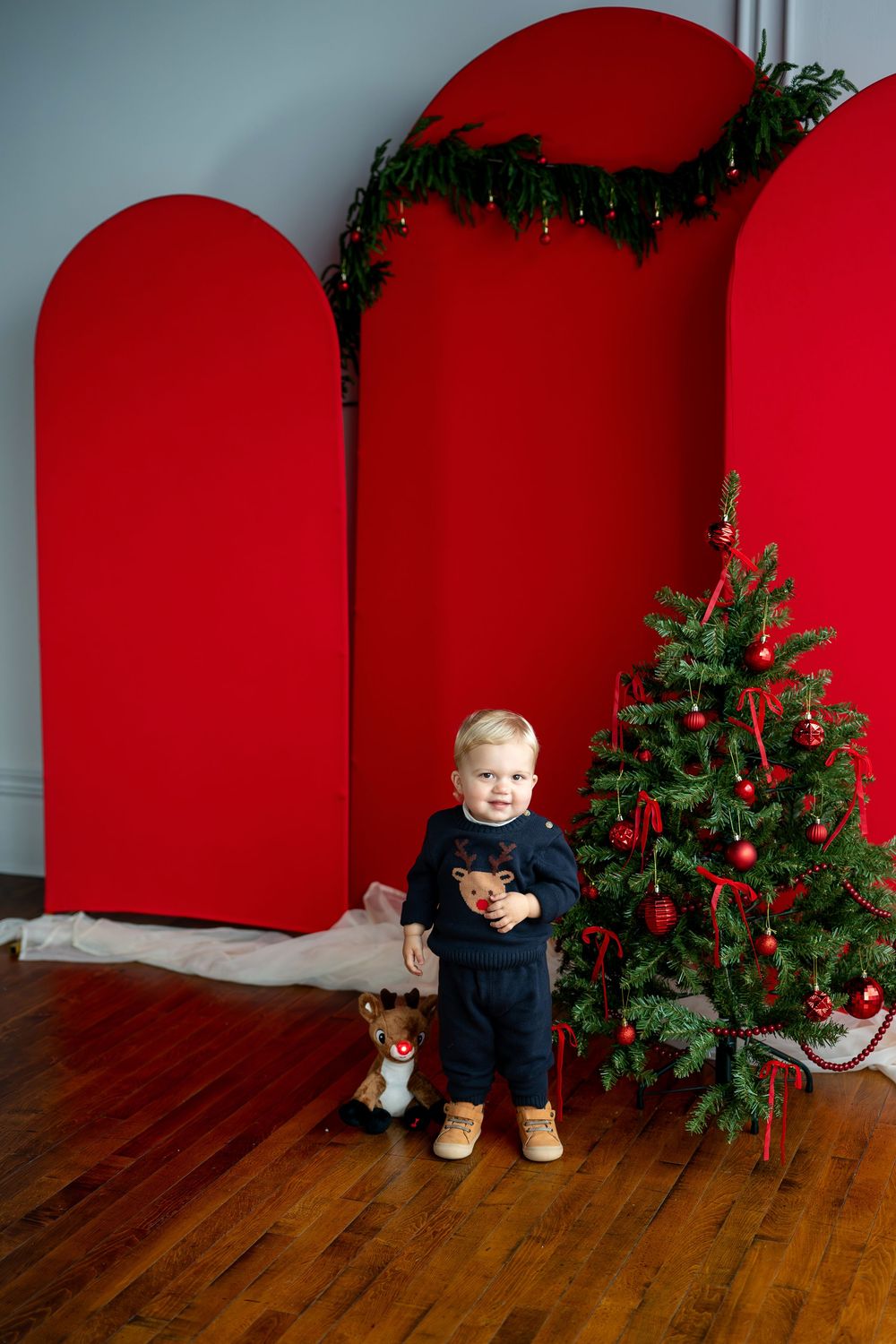 Cute toddler in navy reindeer sweater standing by a Christmas tree with a stuffed reindeer toy nearby.