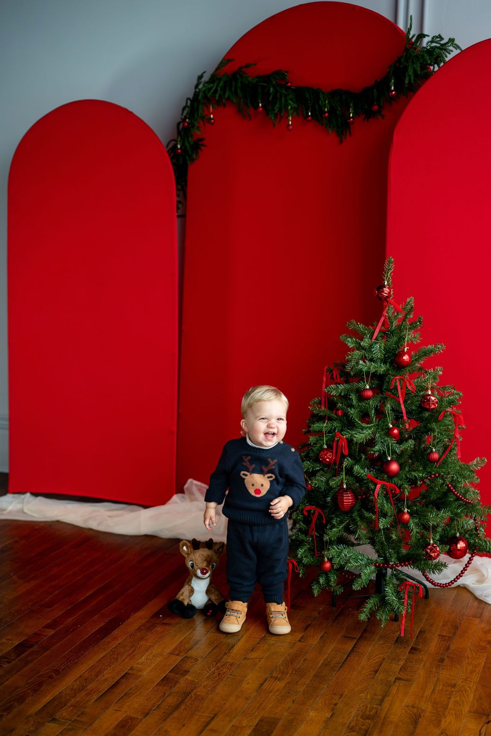 Laughing toddler in navy reindeer outfit standing next to a Christmas tree with red arched backdrop.