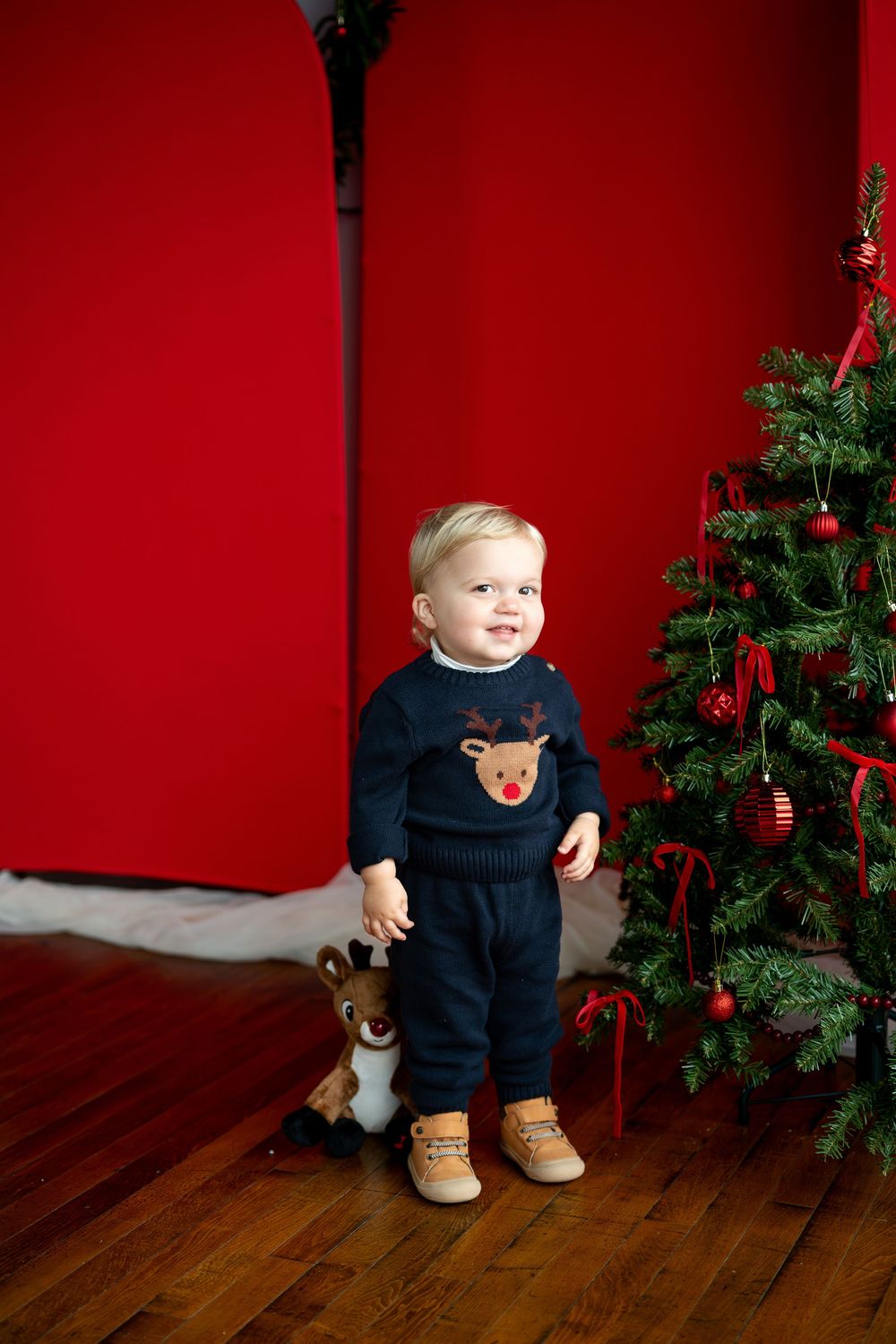 Adorable toddler in navy reindeer sweater smiling beside a Christmas tree with red ornaments and backdrop.