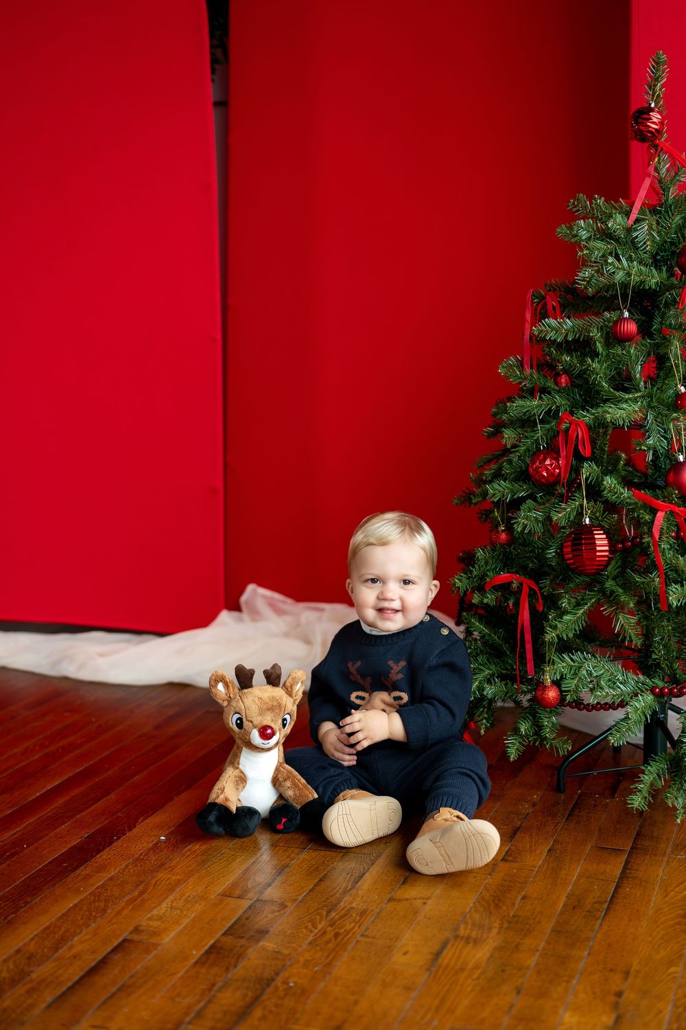 Happy toddler in navy reindeer sweater sitting beside a stuffed Rudolph toy and decorated Christmas tree.