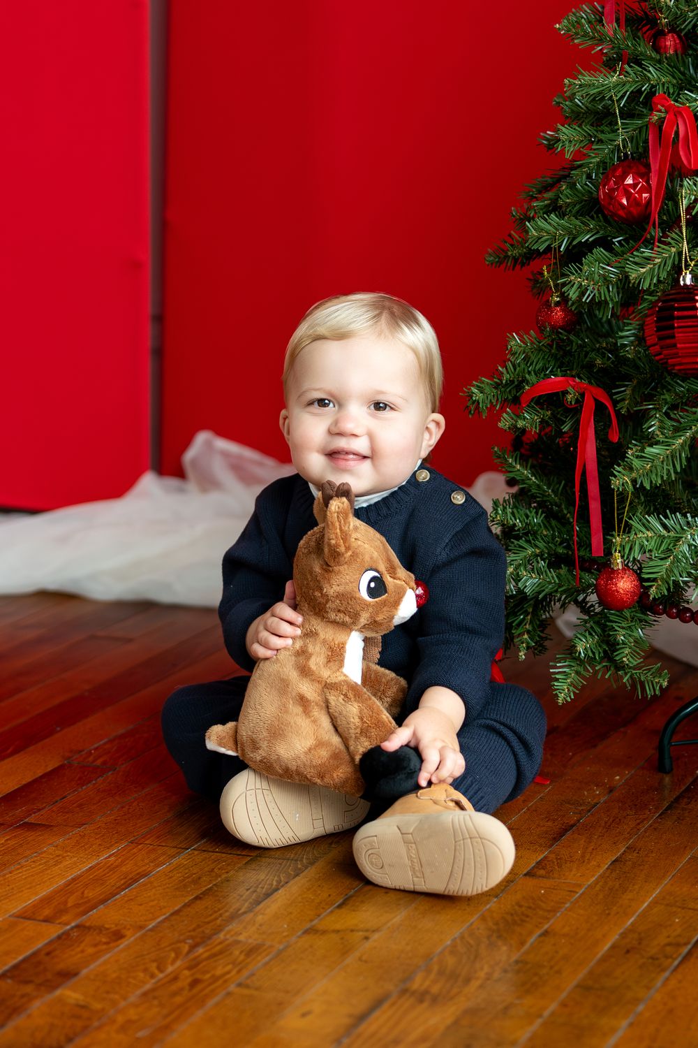 Smiling baby boy in navy outfit holds Rudolph stuffed animal beside decorated Christmas tree on red background.