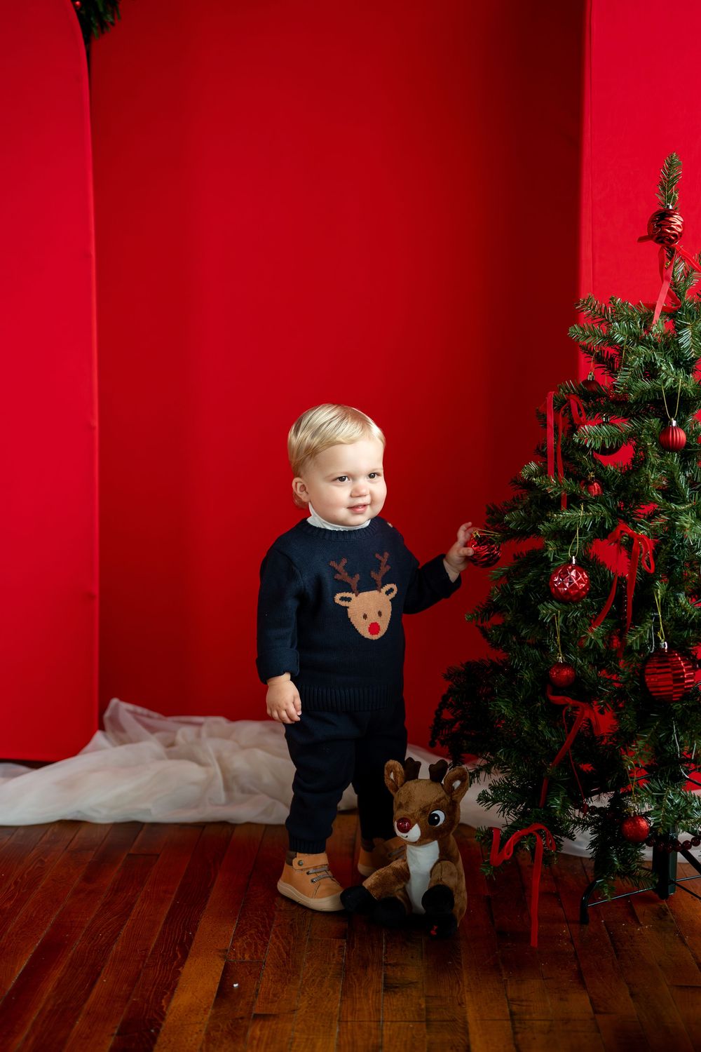 Toddler in reindeer sweater touches Christmas tree ornaments against bright red wall.