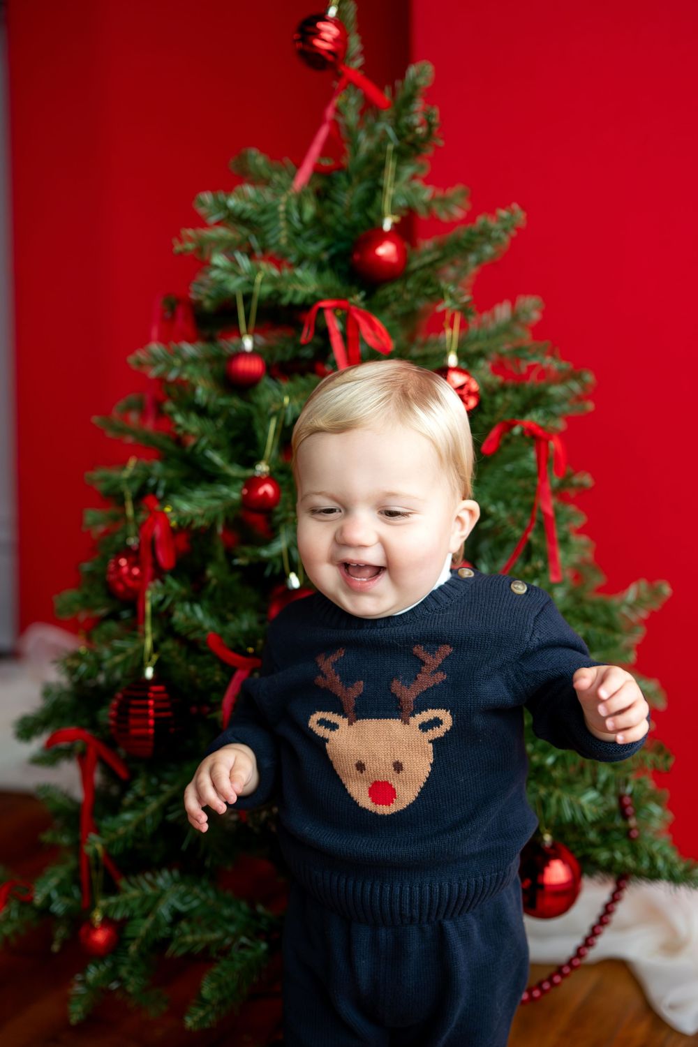 Laughing baby in navy reindeer sweater stands in front of red-ornament decorated Christmas tree.