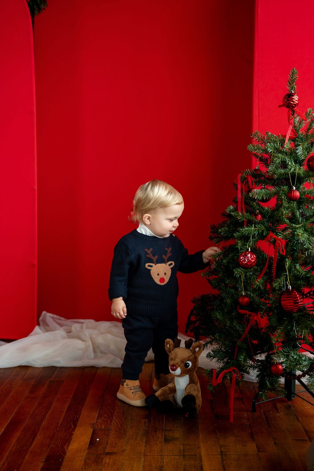 Toddler in navy reindeer outfit reaches for Christmas tree decorations on red background.